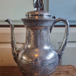 May include: Silver antique coffee pot with a lid and handle. The pot is decorated with grape and leaf patterns. The spout and handle have ornate detailing. The lid is topped with a decorative finial.