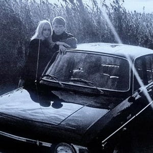 Puede incluir: Fotografía en blanco y negro de un coche antiguo con dos personas apoyadas en el capó. El coche está aparcado cerca de hierba alta. La imagen es en blanco y negro.
