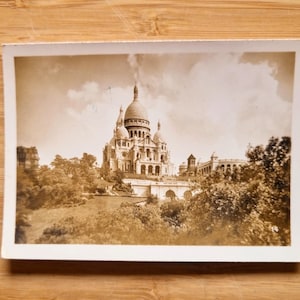 以下が含まれることがあります： フランス、パリのサクレ・クール寺院のセピア色の写真。特徴的な白いドームと複雑な建築様式を持つ寺院は、曇り空を背景にしています。緑豊かな木々が建物を囲んでいます。