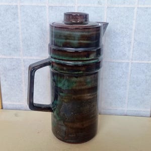 May include: A brown and green ceramic jug with a lid and a handle. The jug has a mottled glaze with brown, green, and black streaks.
