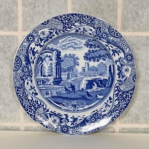 Könnte beinhalten: Ein blauer und weißer Keramikteller mit einem floralen Rand und einem zentralen Bild einer Landschaft mit einer Brücke und einem Gebäude. Der Teller ist in einem traditionellen blau-weißen Muster verziert.