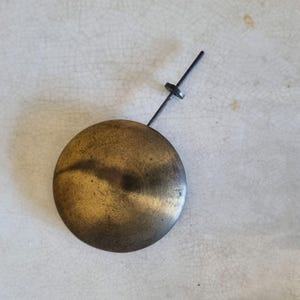 May include: A brass pendulum bob with a black metal rod and a small metal ring at the top.