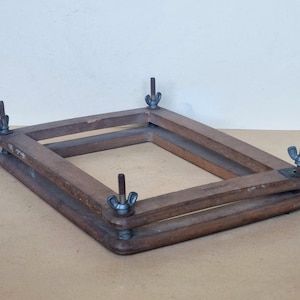 Puede incluir: Un marco de madera con cuatro tuercas de mariposa de metal. El marco está hecho de dos piezas de madera que se unen en las esquinas. El marco está diseñado para sujetar un trozo de tela u otro material.