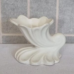 May include: White ceramic vase with a unique, organic shape resembling a seashell or a horn. The vase has a wide, open top and a narrow base.