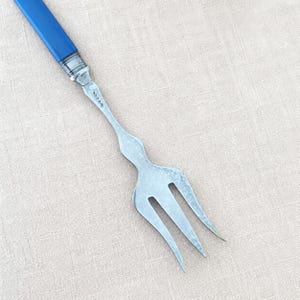 Op de afbeelding: Een zilverkleurige metalen serveervork met een glad, effen blauw handvat. De metalen hals van de vork is voorzien van een reeks kleine, gestempelde keurmerken. De vorkkop heeft drie afzonderlijke tanden met een gebogen, taps toelopend ontwerp.