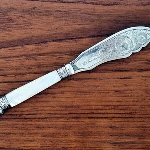 May include: A silver butter knife with an ornate, floral design on the blade. The handle is white with a shell-shaped end. The blade has the word "ecca" engraved on it. The knife is set against a dark wood surface.