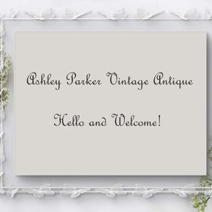May include: A framed sign with the text "Ashley Parker Vintage Antique" and "Hello and Welcome!" in elegant script. The frame is white with butterfly accents, and white flowers are arranged around the frame.