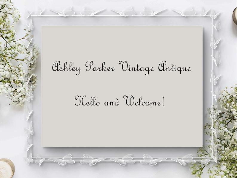 May include: A framed sign with the text "Ashley Parker Vintage Antique" and "Hello and Welcome!" in elegant script. The frame is white with butterfly accents, and white flowers are arranged around the frame.