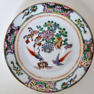 Könnte beinhalten: Eine weiße Porzellansaucers mit goldfarbenem Rand und einem farbenfrohen Blumen- und Vogeldesign. Das Design zeigt rosa, rote, grüne, blaue und gelbe Blumen sowie zwei Vögel mit orangefarbenen und roten Federn.