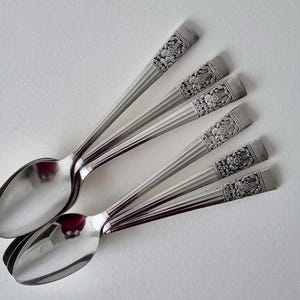 Puede incluir: Seis cucharas plateadas con diseños florales en los mangos, dispuestas sobre una superficie blanca. Las cucharas tienen un acabado brillante y un diseño clásico, ideal para cenas formales. Miden aproximadamente 18 cm de largo.