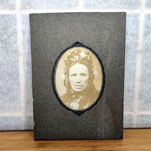Puede incluir: Retrato antiguo en sepia de una mujer en un marco ovalado. La mujer lleva un tocado de encaje. El marco es negro y el fondo es de color gris oscuro. La foto está montada sobre una tarjeta gris.
