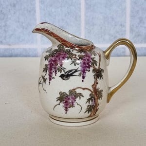 Puede incluir: Una pequeña jarra de cerámica blanca con asa y borde dorados. La jarra está decorada con glicinas moradas y un pájaro negro. El pico tiene un ribete rojo y dorado. Una pieza decorativa.