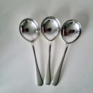 以下が含まれることがあります： 白い表面に配置された、光沢のある銀色のスープおたま3つ。各おたまは丸いボウルと細長い柄を持っています。おたまの反射面は周囲の環境を映し出しています。