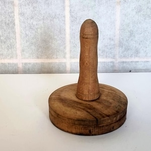 May include: A wooden butter press, featuring a circular base and a long, cylindrical handle. The wood displays a warm, natural colour with visible grain. The press is designed for kitchen use.