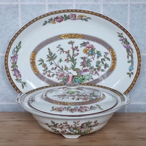 Könnte beinhalten: Eine weiße Porzellanservierschale mit Deckel. Schale und Deckel sind mit einem floralen Muster in Rosa-, Grün- und Gelbtönen verziert. Das Muster zeigt einen stilisierten Baum mit Ästen und Blättern.
