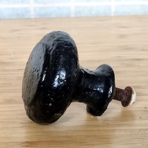 May include: A black, textured cabinet knob with a rounded face and a threaded bolt. The knob has a slightly glossy finish and is lying on a wooden surface. The bolt has a rusty appearance.