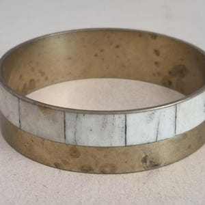 Puede incluir: Una pulsera de brazalete redonda de latón con una incrustación rectangular blanca. La pulsera tiene un aspecto vintage con algo de decoloración. La incrustación blanca está en el medio.