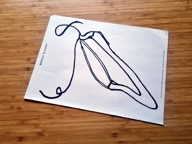 May include: A white sheet of paper with a black line drawing of a shoe. The text "MANOLO BLAHNIK" is printed on the left side of the paper. The paper is on a wooden surface.
