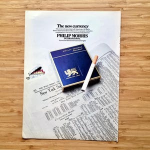 Op de afbeelding: Een vintage advertentie voor Philip Morris International sigaretten. De afbeelding toont een blauw en goud sigarettenpakje, een aangestoken sigaret en een krant. De tekst bevat "The new currency".
