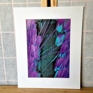 May include: Abstract art print featuring a vibrant composition of purple and teal brushstrokes against a dark background. The artwork is presented within a white mat, creating a striking contrast. The piece is mounted on a wooden surface.
