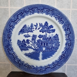 May include: A blue and white porcelain plate with a willow pattern design. The plate features a traditional Chinese landscape scene with a bridge, trees, and a pagoda.