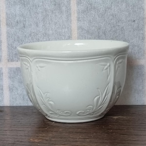 May include: A white ceramic bowl with an embossed floral design. The bowl has a rounded shape and a smooth, white glaze.
