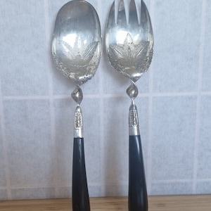 Puede incluir: Un juego de dos cubiertos de servir ensalada de plata con mangos negros. Los cubiertos tienen un diseño floral y un tallo retorcido.