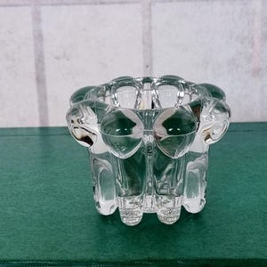 Puede incluir: Un portavelas de cristal transparente. El portavelas tiene una parte superior redondeada con detalles en forma de burbujas y un cuerpo cilíndrico. La base tiene una serie de pequeños pies. El portavelas está sobre una superficie verde.