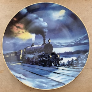 Può includere: Un piatto in porcellana blu e bianca con un bordo dorato presenta un dipinto di un treno a vapore che viaggia attraverso un paesaggio innevato sotto un cielo notturno. Il treno è illuminato da una luna brillante e stelle. Il piatto è firmato "Reg. Lambart".