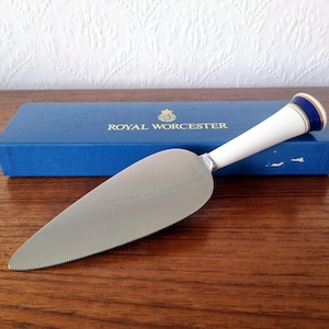 Puede incluir: Una espátula para tartas con borde dentado y mango blanco con detalles azules y dorados. La espátula es de acero inoxidable y descansa sobre una superficie de madera. Una caja azul con texto dorado está en el fondo.