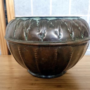 Pode incluir: Uma tigela redonda de metal cor de cobre, com aspeto vintage e pátina escura. A tigela tem uma superfície texturizada com ranhuras verticais e uma borda ligeiramente alargada. A borda tem um tom esverdeado e a tigela assenta numa base escura.