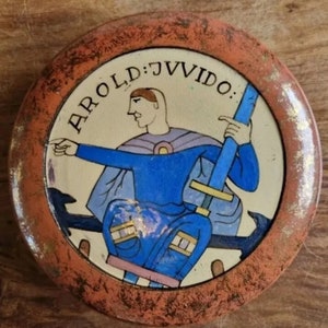 May include: A round ceramic plate with a red and gold border. The centre of the plate features a painted image of a man in blue and purple clothing holding a sword. The text "AROLD: JVVDIO:" is written around the edge of the plate.