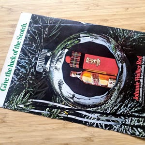 Puede incluir: Un anuncio de Johnnie Walker Red Scotch, que muestra una botella y una caja de regalo dentro de una bola de Navidad. El texto dice "Give the luck of the Scotch" y el nombre de la marca y su eslogan.