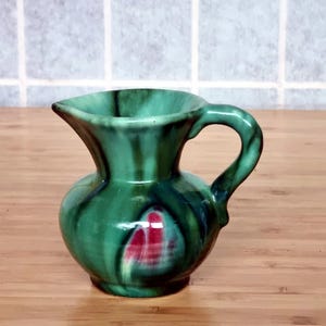 Peut inclure: Un petit pichet en céramique vert avec une anse et un bec verseur. Le pichet a une base bulbeuse et un dessus évasé. Il présente un motif tourbillonnant de vert foncé et d'accents rouges. Le pichet est sur une surface en bois.