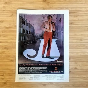 May include: Vintage advertisement featuring a man in a rust-colored suit, light pink shirt, and tie. The background shows a European street scene. The text reads, "For a New World of Fashion... We Search the 'Old World' for Fabric."