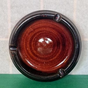 May include: A brown and black ceramic ashtray with a circular design. The ashtray has a deep well and a raised rim.