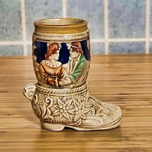 May include: A ceramic boot-shaped mug featuring a detailed design of two people in medieval attire. The mug is decorated with gold, blue, and green accents.