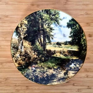 May include: A decorative plate with a gold rim featuring a pastoral landscape scene. The image depicts trees, a stream, and a flock of sheep. The plate is set against a wooden background.