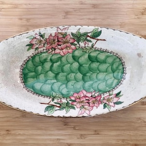 Puede incluir: Un plato ovalado blanco para servir con un diseño floral verde y rosa. El plato tiene un borde festoneado y un borde elevado.