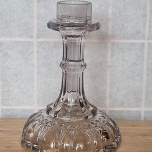 May include: A clear glass candle holder with a fluted design and a scalloped base. The holder is a light purple color.