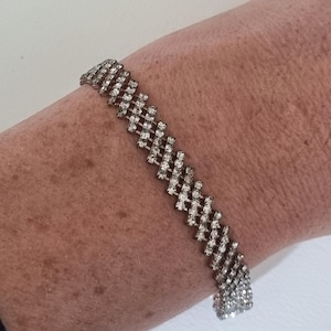 Puede incluir: Una pulsera plateada con un diseño de enrejado, adornada con numerosas piedras de estrás pequeñas y transparentes. La pulsera se lleva en una muñeca, mostrando su apariencia brillante. La joya es un accesorio de moda.