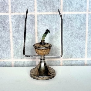 May include: A vintage silver-toned oil lamp with a decorative base and a metal frame. The lamp features a glass reservoir and a wick. The lamp is designed to hold oil and provide light.