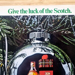 Puede incluir: Un anuncio con una bola de Navidad de cristal transparente con una botella de whisky escoc&eacute;s y una caja de regalo en su interior. La bola cuelga frente a un fondo de agujas de pino verdes. El texto "Give the luck of the Scotch" est&aacute; en la parte superior.