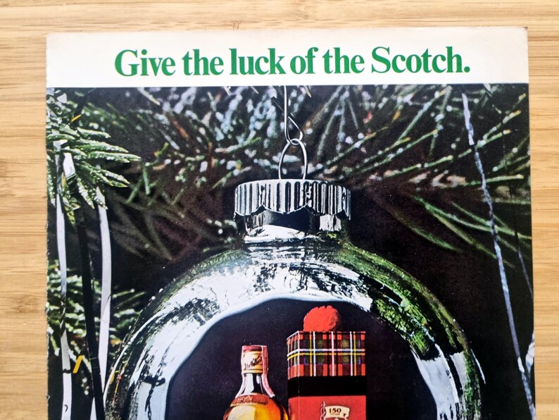 Puede incluir: Un anuncio con una bola de Navidad de cristal transparente con una botella de whisky escoc&eacute;s y una caja de regalo en su interior. La bola cuelga frente a un fondo de agujas de pino verdes. El texto "Give the luck of the Scotch" est&aacute; en la parte superior.
