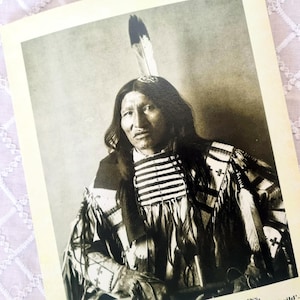 Puede incluir: Retrato en tonos sepia de un nativo americano. Lleva un tocado de plumas y una camisa estampada con rayas horizontales. La imagen es una fotografía antigua.