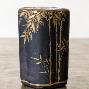 Könnte beinhalten: Eine schwarze, rechteckige Vase mit einem goldenen Bambusdesign. Die Vase hat einen goldenen Rand oben und unten. Die Bambusstängel und Blätter sind in Gold gemalt und bilden einen auffälligen Kontrast zum dunklen Hintergrund.