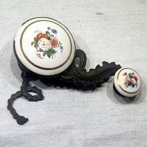 May include: An antique bicycle bell with a white ceramic face featuring a floral design in red, pink, and blue. The bell has a dark metal base and a small matching knob. The bell is attached to a chain.