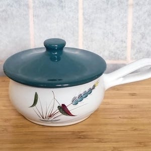 Puede incluir: Una olla de sopa de cerámica blanca con tapa y asa color teal. La olla presenta un diseño floral con detalles en verde, rojo y azul. La olla está sobre una superficie de madera.
