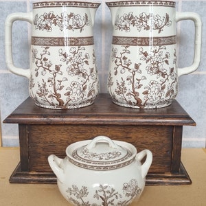 May include: Two white ceramic jugs with brown floral designs and a matching white ceramic sugar bowl with a brown floral design. The jugs and sugar bowl are sitting on a wooden box.
