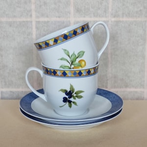 May include: Two white ceramic teacups with blue and gold trim and floral designs. The cups are stacked on top of each other and are sitting on a white saucer with a blue rim.